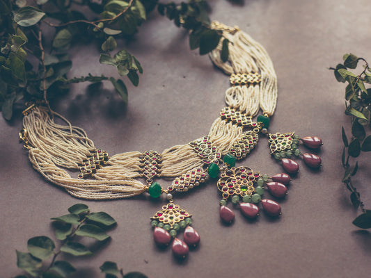Abharika necklace displayed flat showing multi-strand pearl design with ornate kemp stone pendants and pink rhodonite beads, styled with green foliage on textured surface