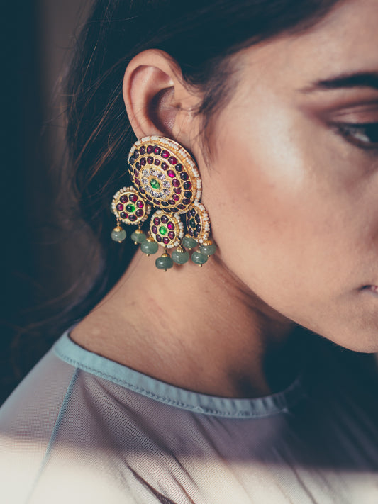 Model wearing Dhanya earrings featuring ornate gold-plated circular motifs with multicolored kemp stones in red, adorned with dangling green bead drops - Aaharya temple jewelry