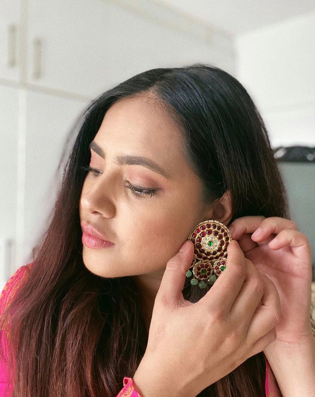 Close-up of Fashion influencer adjusting Dhanya earrings showing detailed circular kemp stone work in red and green with gold-plated finish and green bead drops - Aaharya temple jewelry